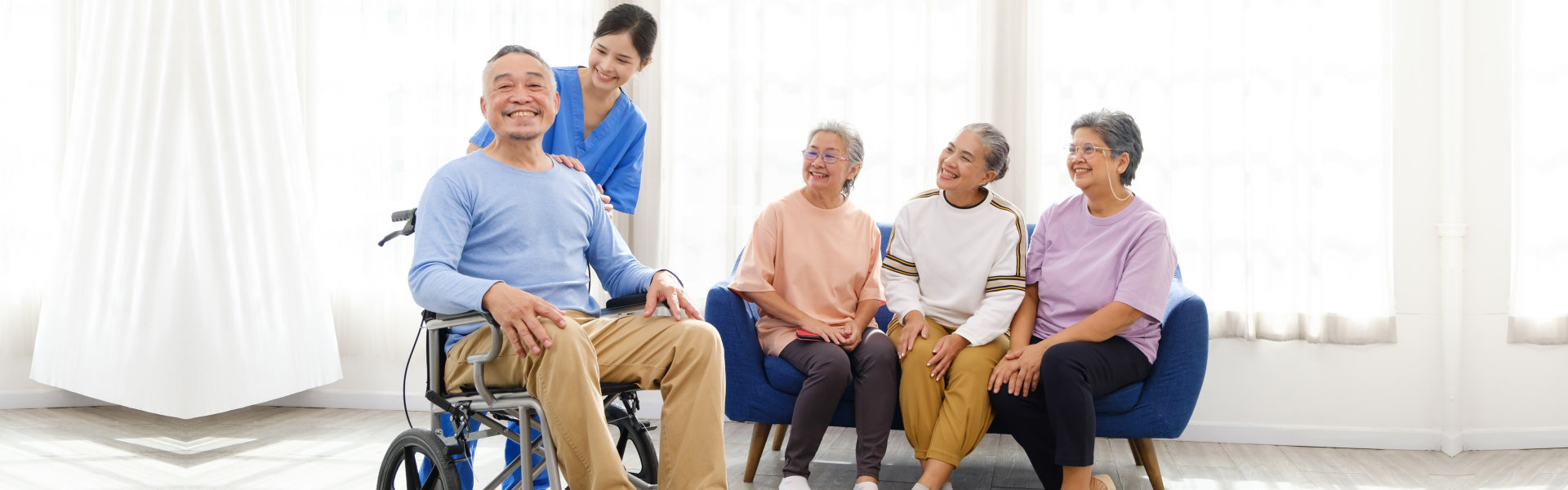 The caregiver therapist stands with a man sitting in a wheelchair with a group of women sitting on a sofa